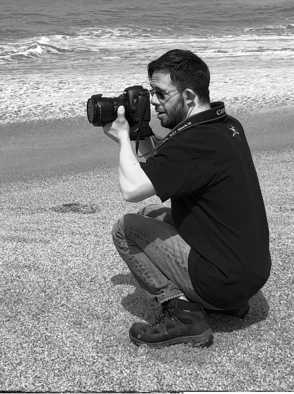 Carlos, en cuclillas sobre la arena, encuadra con su cámara frente al mar.